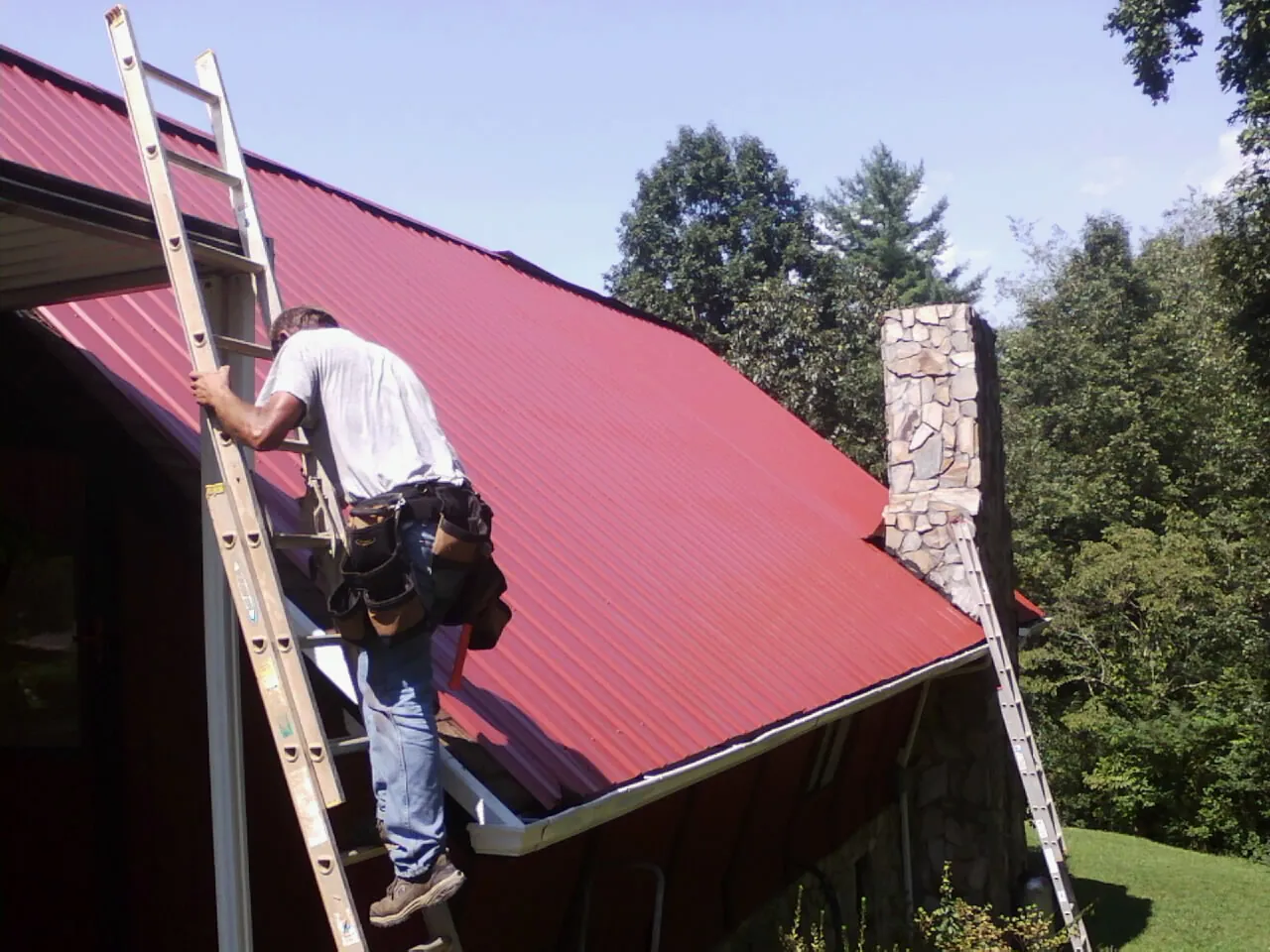 Roofer on a ladder beside a red metal roof and stone chimney — Soffit & Fascia Repair by Roofing Brothers in Hartline
